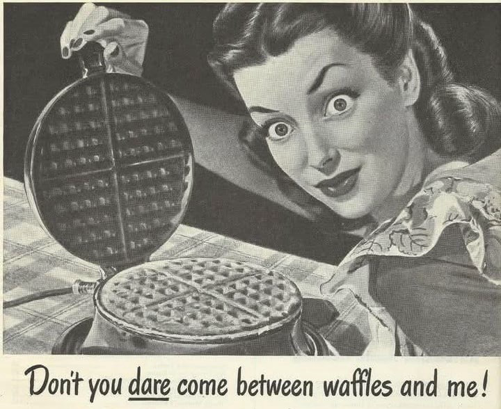 Retro ad. A woman with insane eyes looks directly at you. She’s making waffles with a waffle maker grill thing. A caption reads, “Don't you dare come between waffles and me!”